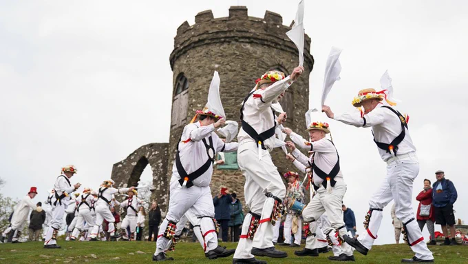 Morris dancers from the Leicester Morrismen perform a traditional May Day dance at dawn in Bradgate Park, wearing white costumes with bells and waving handkerchiefs, with the Old John tower in the background and spectators watching.