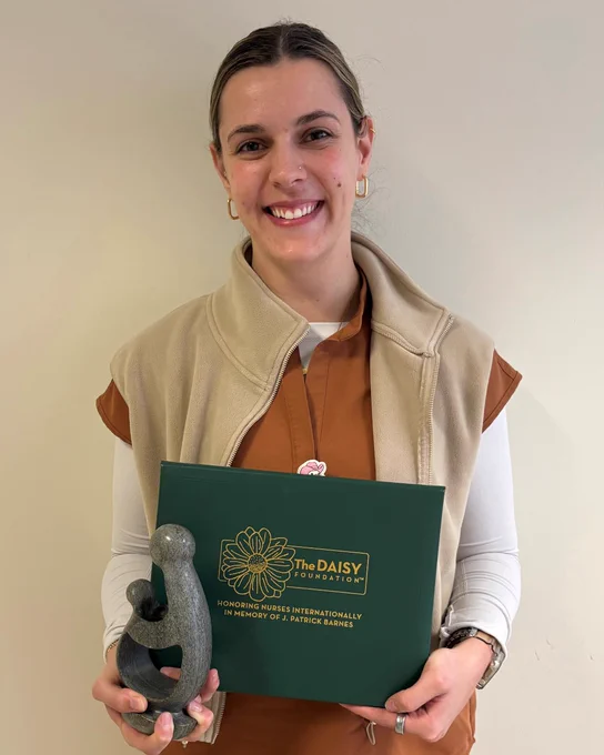 Person holding The DAISY Foundation award plaque and a sculpted trophy against a plain background.