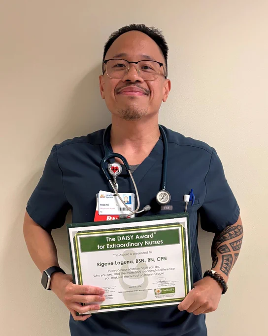 Nurse in navy scrubs holding The DAISY Award certificate for Extraordinary Nurses against a plain wall.