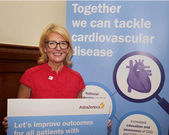 Woman in red holding a sign advocating improved outcomes for cardiovascular disease patients, standing by a banner promoting tackling cardiovascular disease together.
