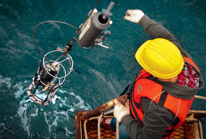 A colour photograph of a man in hard hat and life vest operating machinery on an offshore platform above ocean waters.