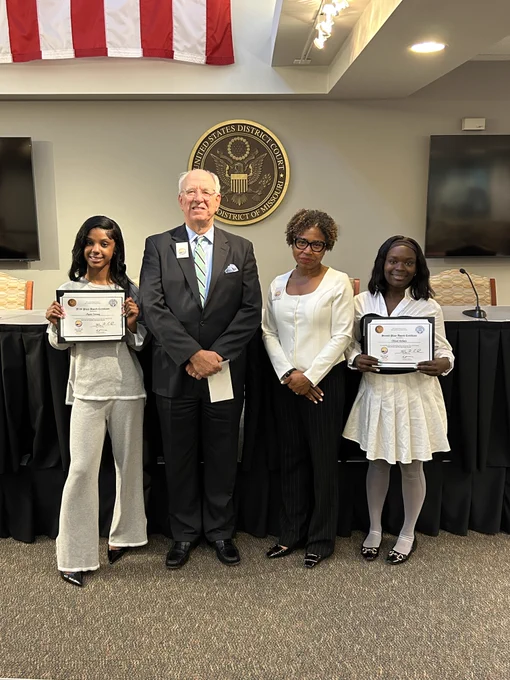 A photo of two students who are winners of the video contest. The students are holding their prize certificates. In between them is JLC Board President Mr. Glenn Davis and U.S. Magistrate Judge Shirley Mensah. Behind them, the circular seal of the U.S. District Court Eastern District of Missouri hangs on the wall
