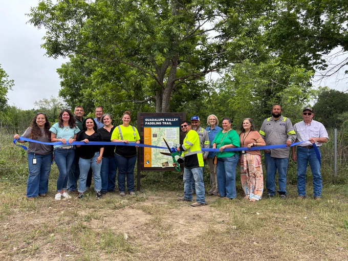 Group of people cut ribbon with oversized scissors next to kiosk that says Guadalupe Valley Paddling Trail