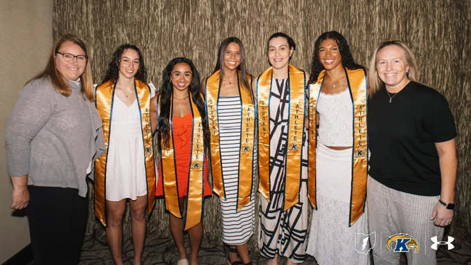 Five Kent State student-athletes wearing gold "Student-Athlete" graduation stoles pose for a group photo with two coaches at a Kent State Athletics recognition event. The group stands in front of a textured neutral backdrop, smiling. The Kent State logo and Under Armour logo appear in the lower right corner of the image.