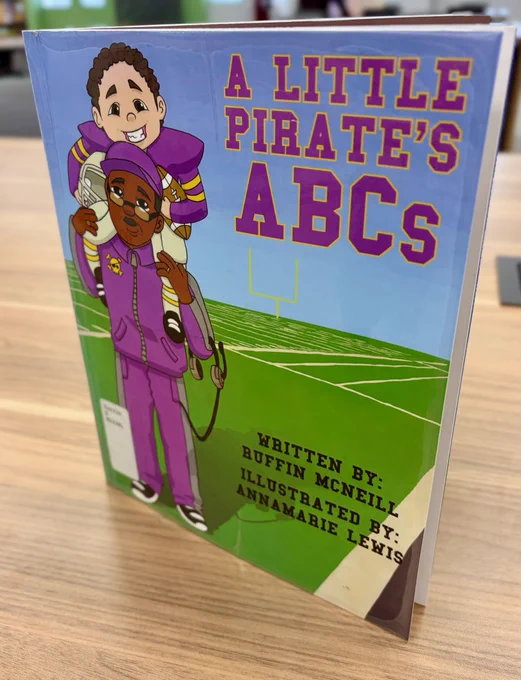 Cover of the children's book titled "A Little Pirate's ABCs" featuring a character in a purple sports uniform holding a young child on their shoulders. The background shows a football field. The book is written by Ruffin McNeil and illustrated by Annamarie Lewis.