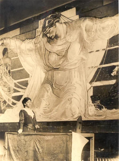 A sepia-toned photo of a woman standing in front of a large tapestry depicting a kneeling, robed figure with outstretched arms.