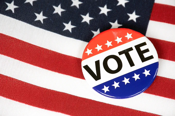 A pin with "VOTE" in black lettering featuring a red top with white stars, white center, and a blue bottom with white stars, against an American flag background.