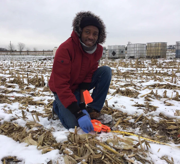 Dr. Emmanuel Badewa setting up a winter field experiment to quantify greenhouse gas emissions from biobased amendments during his doctoral research at the University of Waterloo. Credit: Nadia Gabbanelli.  ALT A man wearing a hooded winter jacket and gloves holding a measuring tape in a farm field covered with corn stubble and snow