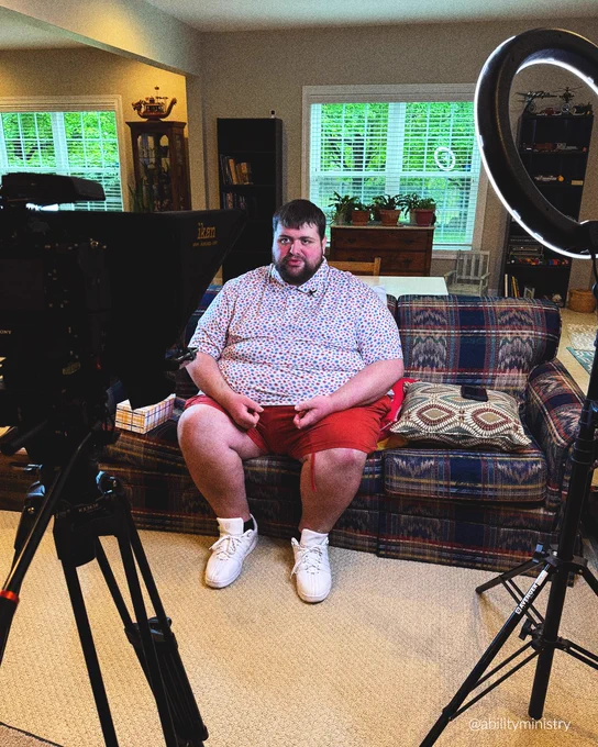 A young adult man is seated on a couch surrounded by recording equipment, as he reads a disability ministry curriculum lesson ion camera.