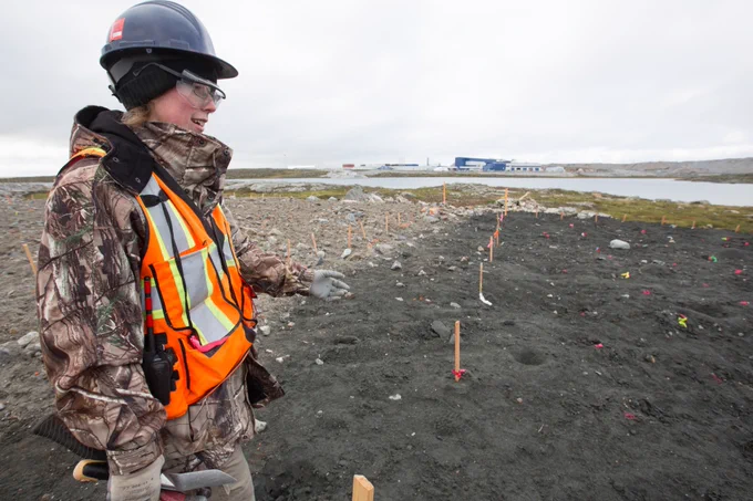 A person wearing a hard hat and a safety vest and goggles standing over new soil. Orange markets are set a few meters apart.