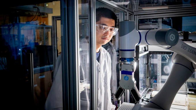Scientist in a lab coat operating a robotic arm inside a glass-enclosed laboratory workspace.