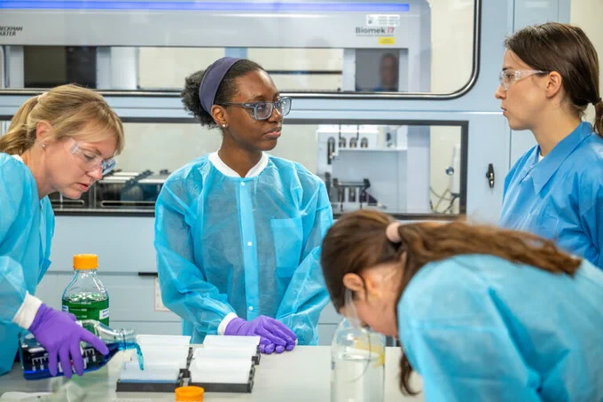 Four scientists in blue lab coats and purple gloves working together in a laboratory setting with scientific equipment on the table.