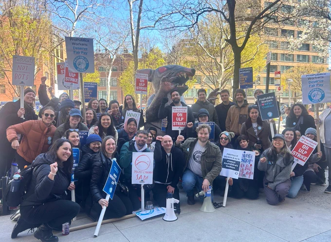 A group of about 40 people stand or squat on a sidewalk. Many have fists raised in the air. Many are hodling signs that read "ON ULP STRIKE", "PROPUBLICA WORKERS DESERVE FAIR PAY", "THOUGHTS NOT BOTS", and "PROPUBLICA WORKERS HEART A FAIR CONTRACT". 