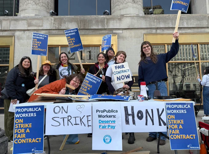A group of about 8 people stand on the sidewalk in front of a table covered in signs. The signs say "PROPUBLICA WORKERS ON STRIKE FOR A FAIR CONTRACT" and "HONK". The people are holding signs and smiling. 