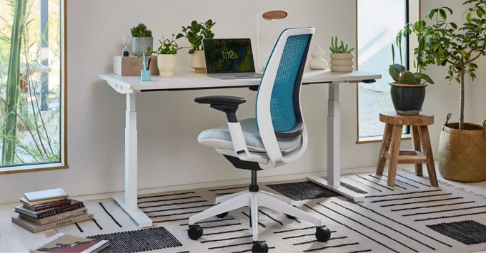 A Steelcase Amia Office Chair with blue mesh back at a white adjustable desk in a bright room with plants and natural light.