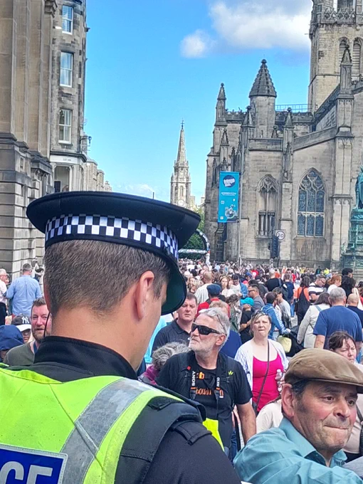 A police officer surveys a large crowd gathered on a street with historic buildings and a church spire.