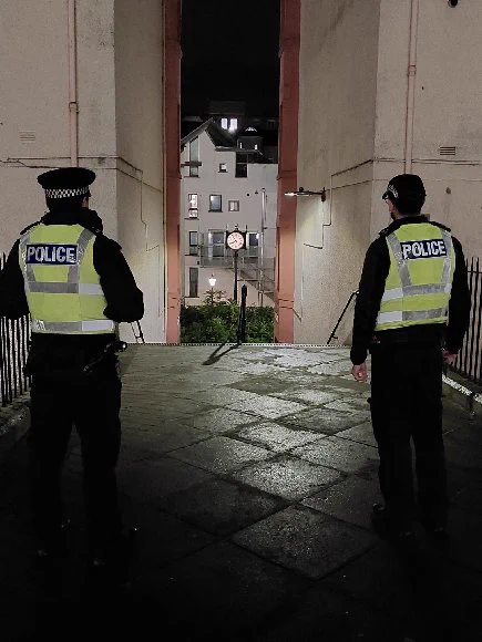 Two police officers in high-visibility vests stand in a wet courtyard at night, with a modern building in the background.