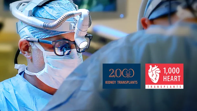 A surgeon wearing protective eyewear and a surgical cap operates in a well-lit room. Next to the surgeon, logos display "2,000 Kidney Transplants" and "1,000 Heart Transplants.