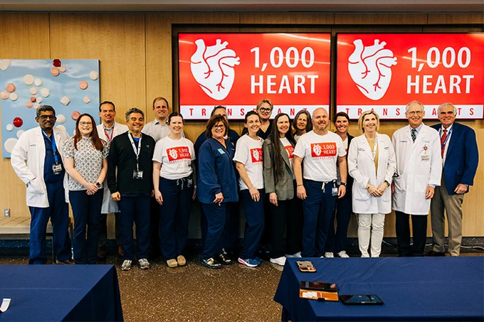 A group of people, dressed in a mix of casual and professional attire, stand in front of two screens displaying a heart graphic with the text "1,000 Heart Transplants." They appear to be celebrating a milestone in heart transplants.