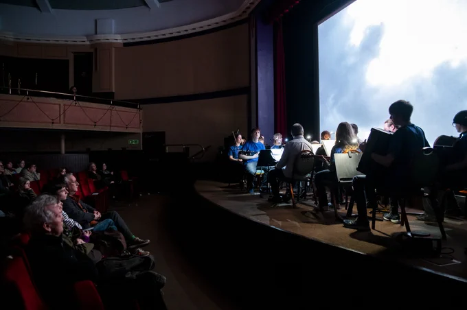 A dimly lit audience is visible watching an ensemble of young musicians and a cinema screen.
