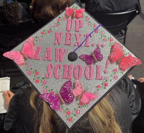 Close-up of a decorated graduation cap featuring glitter and pink butterflies with the message “Up Next: Law School.” 