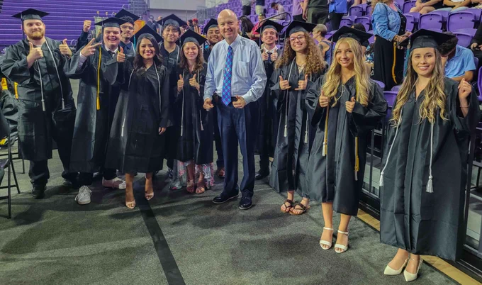 Smiling group of graduates in caps and gowns giving thumbs-up gestures while standing with Dr. Jeff Allbritten on the arena floor, with audience members and purple stadium seating in the background. 