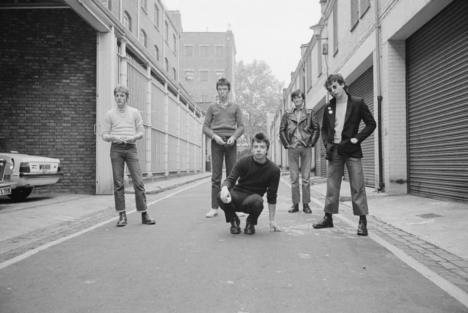 Black-and-white street promo shot of The Undertones, circa 1979. The band stand spaced across a narrow urban road, relaxed but locked in presence — a raw, grounded image that mirrors the sharp, fast-paced energy of their debut era.