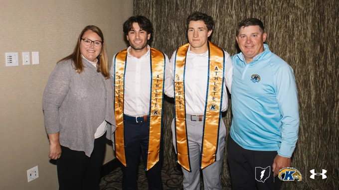 Kent State men's golf graduation photo with James Newton (middle left) and Ben MacLean (middle right) standing between Associate Athletic Director for Student-Athlete Support Services Amy Densevich on their left and head coach Jon Mills on their right. James and Ben, both smiling, wear white dress shirts with gold "Student Athlete" graduation stoles featuring the Kent State logo — James in navy pants with a brown belt, Ben in gray pants with a tan belt. Amy wears glasses and a gray sweater with black pants. Jon wears a light blue Kent State Golf quarter-zip pullover with dark pants. They're posed in front of a textured brown curtain backdrop. Ohio Sports Lightning, Kent State, and Under Armour logos are in the bottom right.