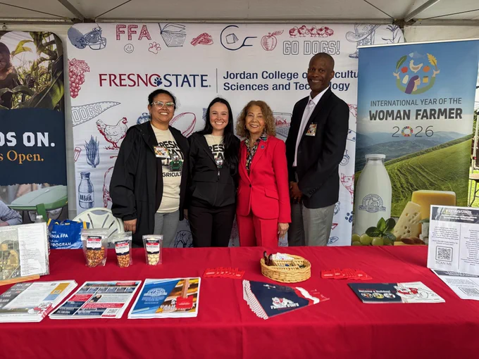 This image shows a group of four people standing under a tent at an event. They are in front of displays for Fresno State's Jordan College of Agricultural Sciences and Technology and the International Year of the Woman Farmer 2026. The table is covered with pamphlets, brochures, and a basket. Banners and posters with related imagery are visible in the background.