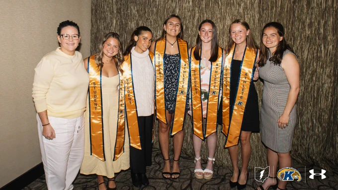 A group of seven people smiling and standing together, wearing sashes with the word "Student-Athlete" and Kent State University insignia. They are in front of a textured wall with Kent State University and Under Armour logos at the bottom.