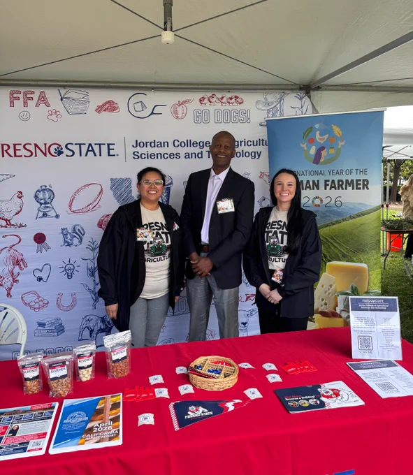 A group of three people standing behind a table adorned with promotional items and a Fresno State banner at a booth. The backdrop features logos and agricultural imagery. A sign highlights "International Year of the Human Farmer 2026.