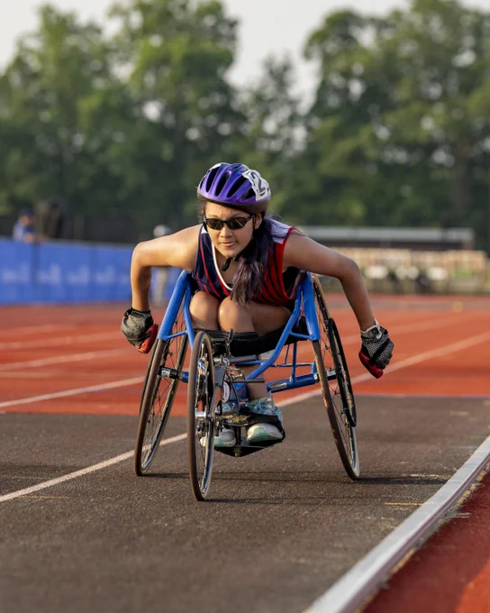 A female athlete in a racing wheelchair on a track, wearing a helmet and sunglasses, pushing forward with determination.