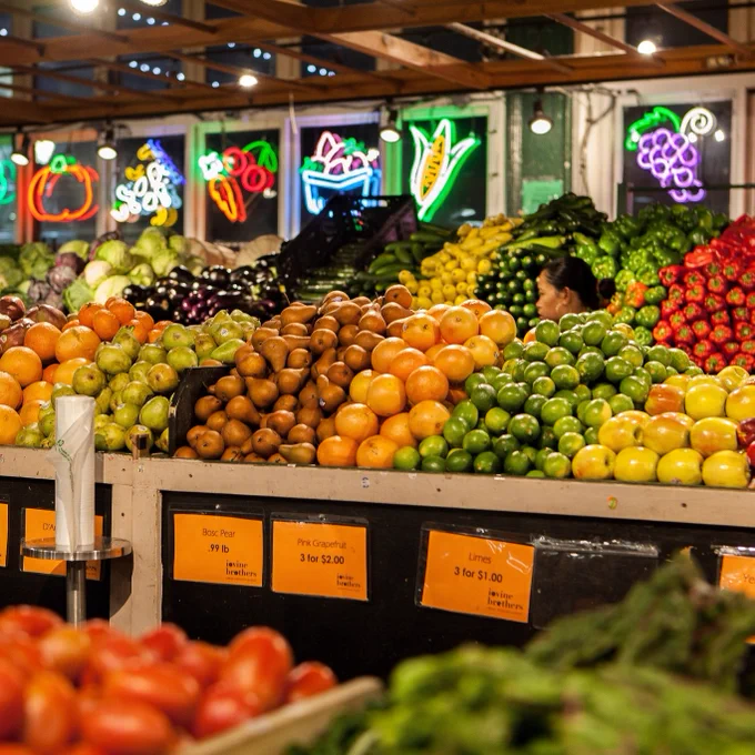 A vibrant display of colorful ripe fruits and vegetables with neon depictions of them behind the display stands