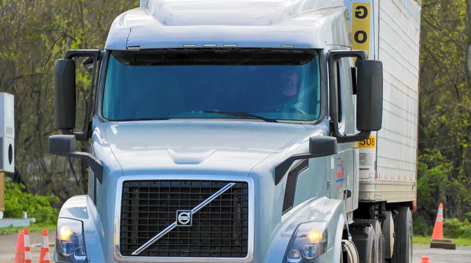 Front view of a silver Volvo semi-truck with a large trailer driving on a road lined with orange traffic cones.