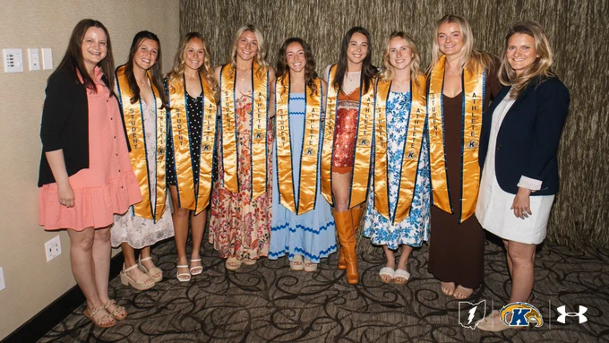 A group of individuals is posing together indoors, each wearing a golden sash. They are dressed in a variety of formal and semi-formal outfits. The image includes logos of organizations including Kent State and Under Armour.