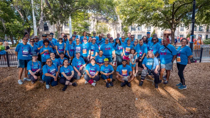 Large group of diverse people wearing matching blue event t-shirts posing outdoors in a park with trees and buildings in the background.