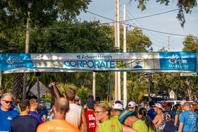 Runners gather at the starting line of the AdventHealth Corporate 5K race under a blue banner on a sunny day.