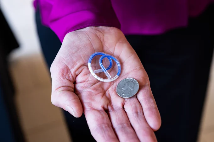 Close-up of a woman's hand holding a pretzel-shaped device next to a quarter for size comparison. 