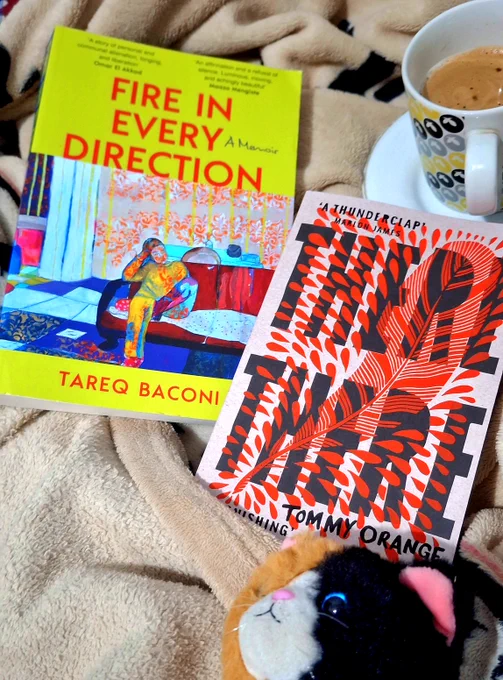 A photo of two books on a beige blanket, flanked by a coffee cup and a plush cat. One is Fire in Every Direction: A Memoir by Tareq Baconi, with a yellow cover and a painting of a man sitting on a red couch. The other is There There by Tommy Orange, with a cover art of a heavily stylized red feathers surrounded by exploding shapes that resemble blood drops.