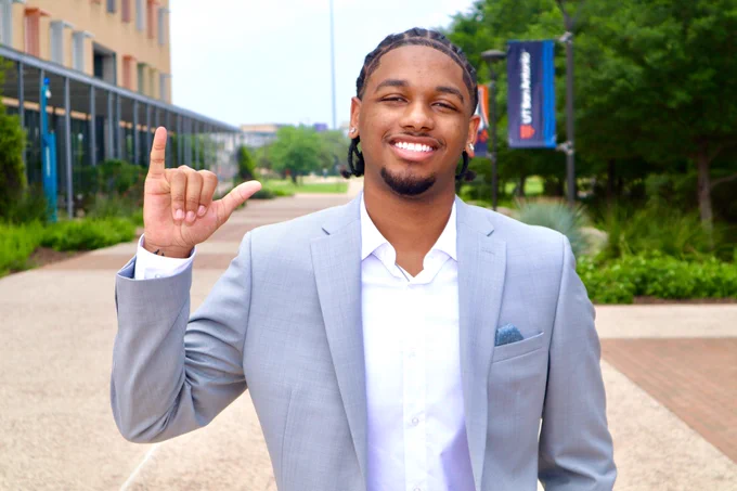 Torey Whitfield in a suit, signing "Birds Up" with his right hand on the UT San Antonio campus, smiling.