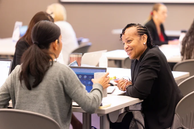 A group of people seated at tables engaged in conversation. Two individuals in the foreground are talking, one smiling, at the 2026 School Finance Conference. Laptops and papers are visible on the tables.