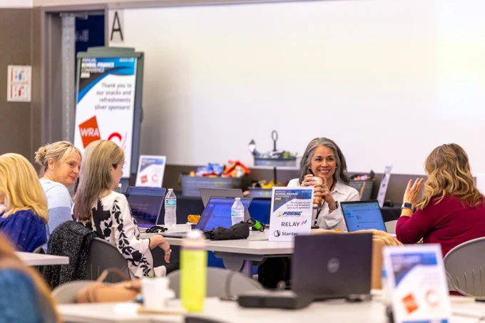A group of people sitting at tables during a professional event. Laptops and papers are on the tables, and a banner with the event sponsor is visible in the background.