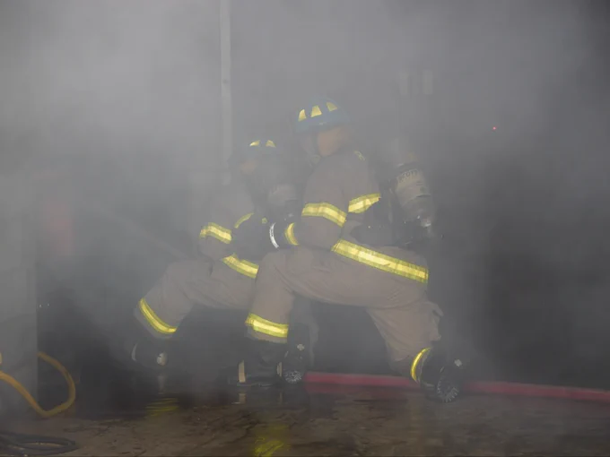 Student firefighters in protective gear navigate a smoke-filled area, wearing helmets and oxygen tanks. They are holding a fire hose, surrounded by dense smoke.