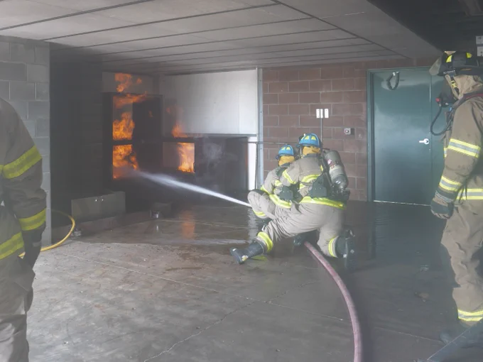 Student firefighters are training in a controlled environment, using a hose to extinguish a fire in a building. The flames are visible, and another firefighter is observing closely. All are wearing protective gear.
