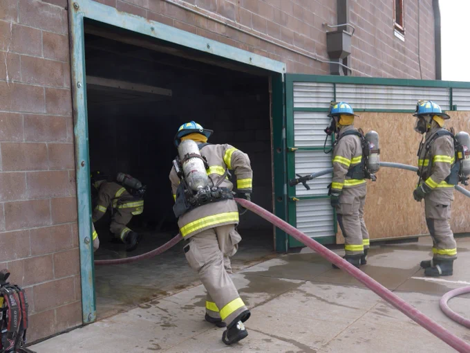 Student firefighters wearing protective gear and helmets hold a hose as they enter a smoky building, preparing to combat a fire. The scene depicts a training or emergency response scenario. Walls are brick, and there's a garage-like door partially open.