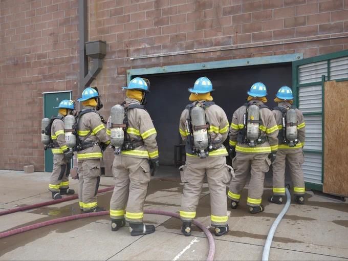 A group of student firefighters, wearing protective gear and helmets, stand outside a building with a smoky doorway. They are equipped with oxygen tanks, and several hoses are on the ground. The firefighters appear ready to enter the building.