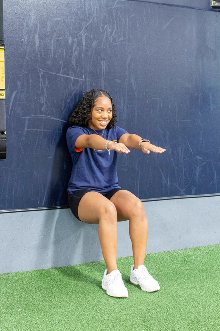 Student doing a wall sit with arms forward and smiling.