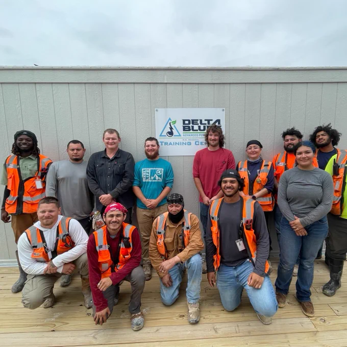 A group of people standing in front of a building with a sign that reads "Delta Blue Training Center." They are wearing various clothing, including orange safety vests.