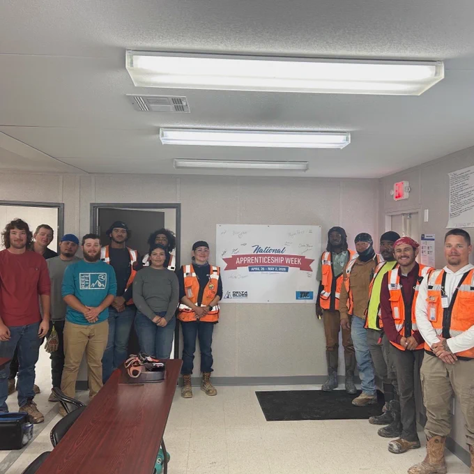 A group of people standing in a room, some wearing orange safety vests. They are gathered near a wall sign that reads "National Apprenticeship Week." Tables and chairs are visible to the side.