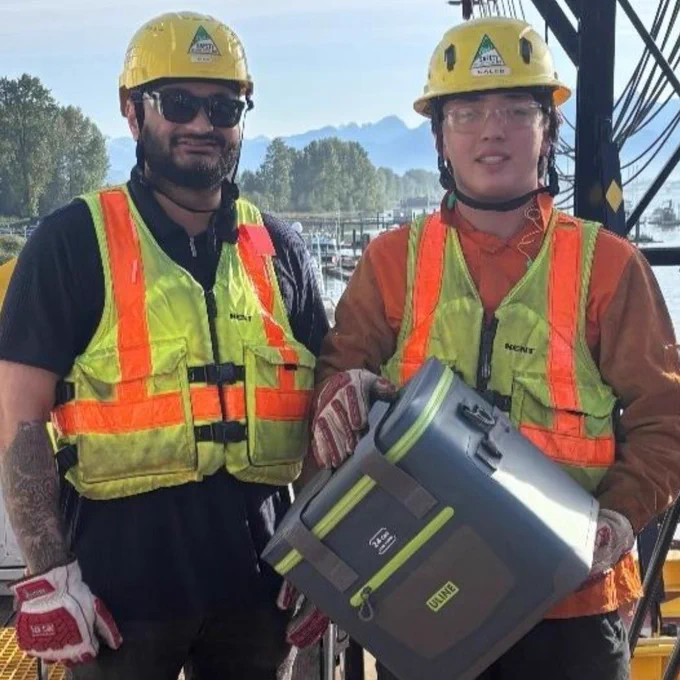 Two construction workers wearing safety gear, including helmets and reflective vests, are standing on a platform. One of them is holding a black and green equipment case. The background features a water body, forested area, and mountains.